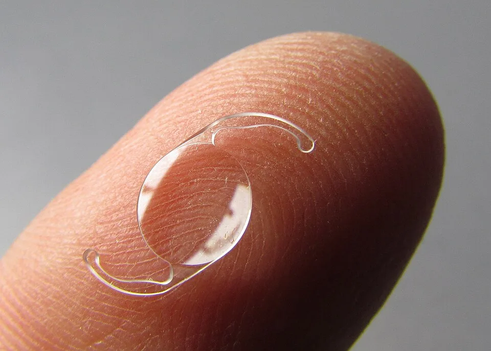 Uma lente intraocular transparente, com duas hastes curvas, apoiada na ponta de um dedo
